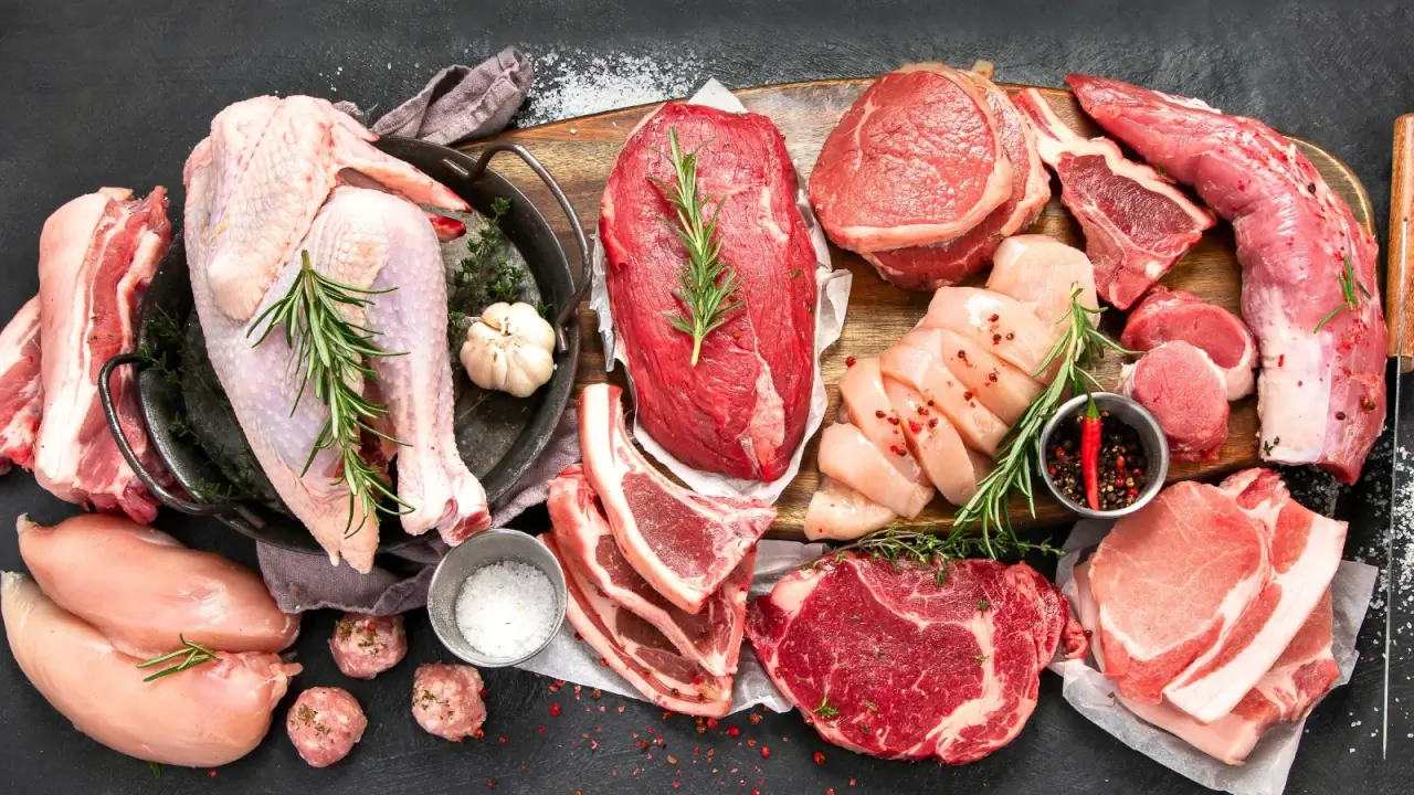 Top view of various types of raw meat cut into pieces, including parts of beef, pork, lamb and chicken with sprigs of green rosemary, heads of garlic, and other spices on a black background.